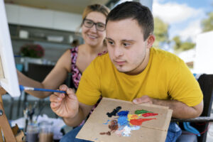 Teen with Down syndrome uses a palette to paint on canvas. His art therapist smiles in the background.
