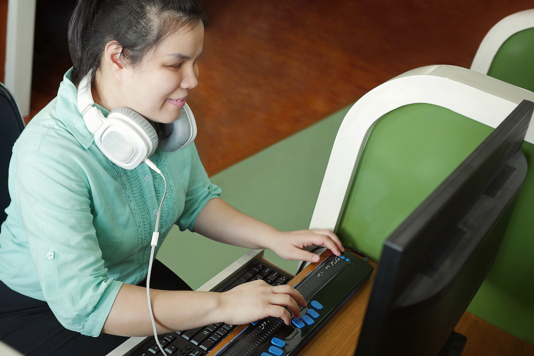 A photo of a blind woman with headphone uses computer with a braille terminal, a technology device for persons with visual disabilities.