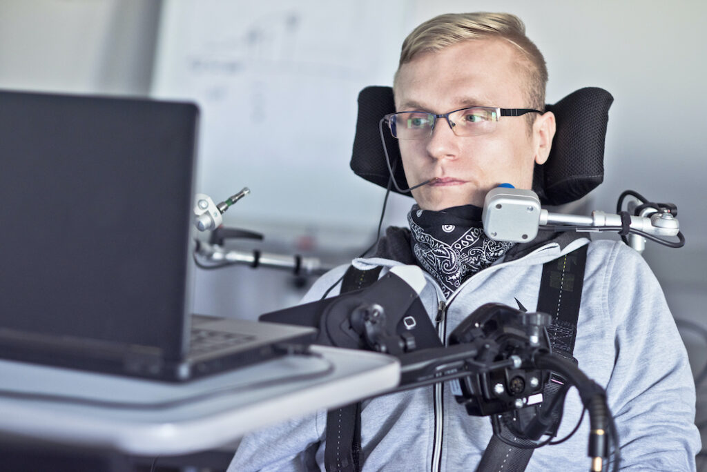 A photo of a disabled user with a lot of personal assistive technology uses a laptop computer.