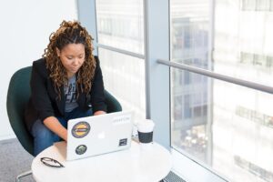 A photo of an artist writes copy for her website on a laptop with a coffee beside a big skyscraper window.