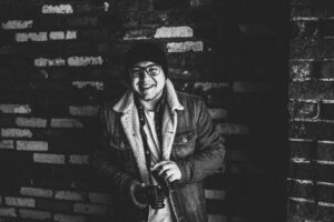 A photo of a smiling photographer with a toque and glasses holds his camera in front of a brick wall.